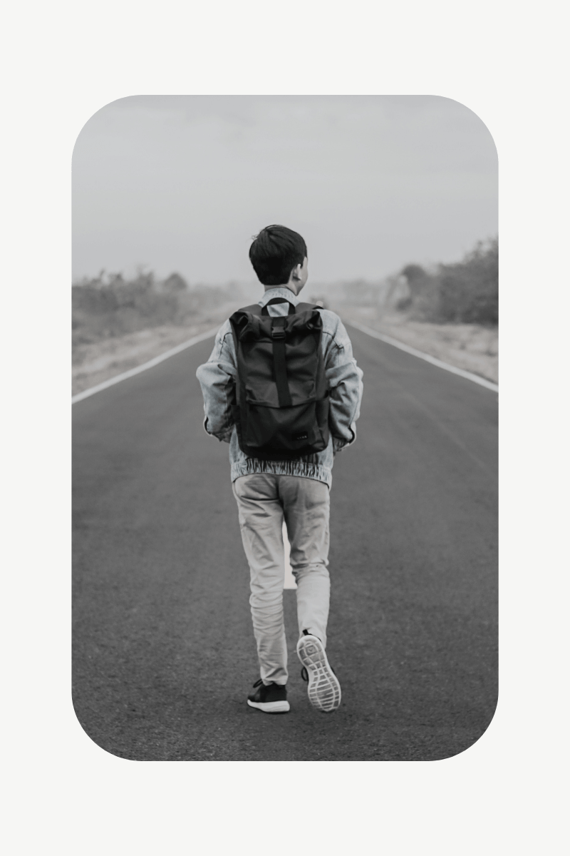 Artistic, high-contrast photograph of a man with a backpack walking down a remote road bordered by desert brush, representing a personal journey and intentional living.